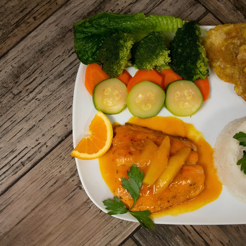 Colombian meal with fish in mango sauce, rice, plantains, and steamed vegetables on a white plate.