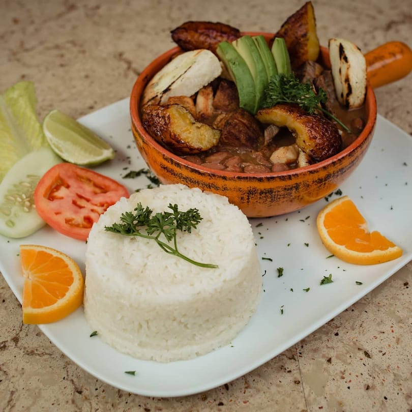 Colombian dish with rice, beans, plantains, avocado, and fresh vegetables on a white plate.