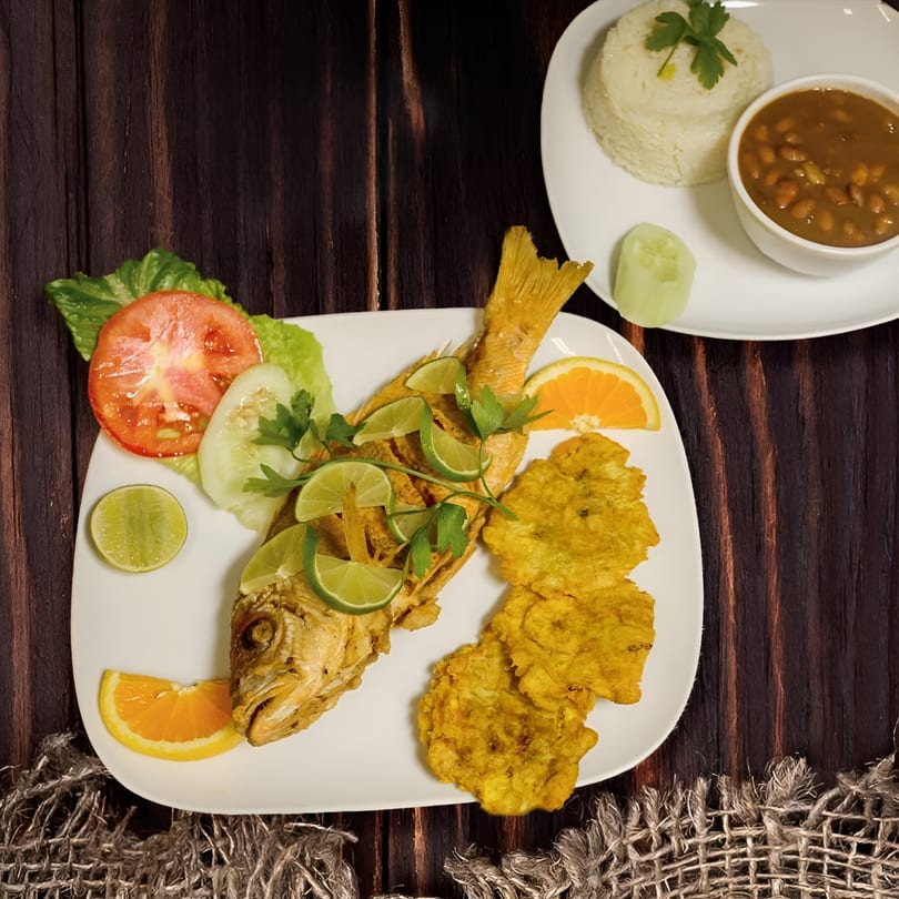 Fried fish with limes, plantains, and salad, with rice and beans in the background.
