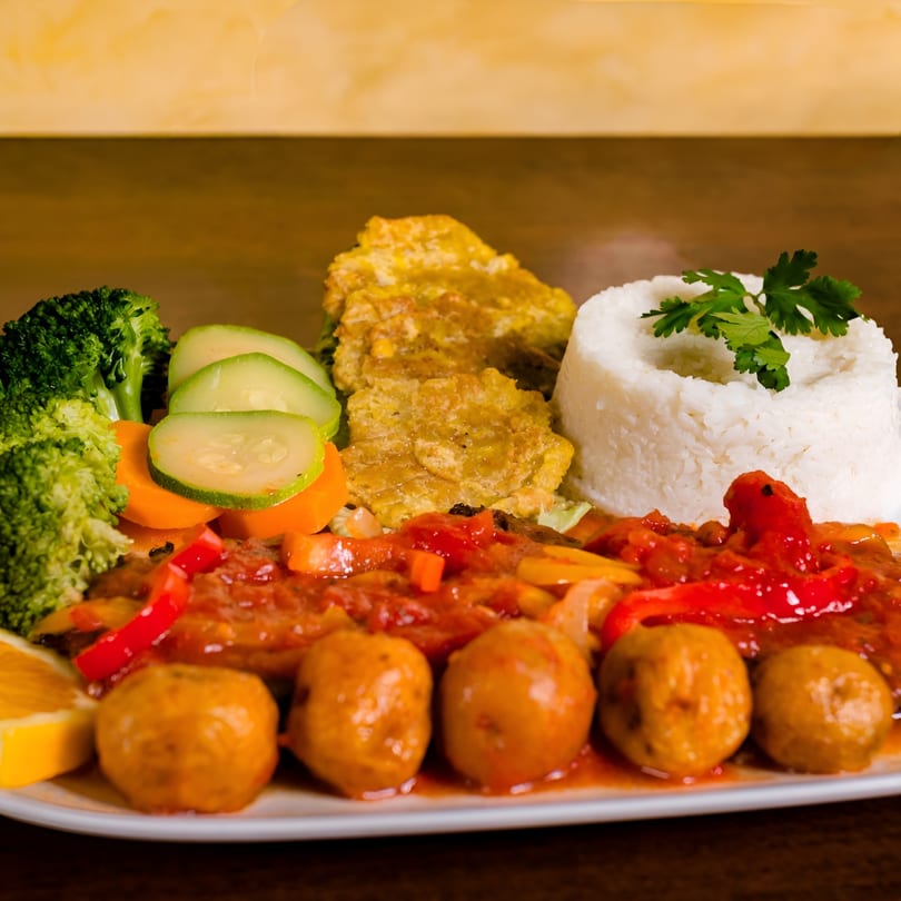 Colombian dish with meatballs in red sauce, rice, plantains, broccoli, and sliced vegetables.
