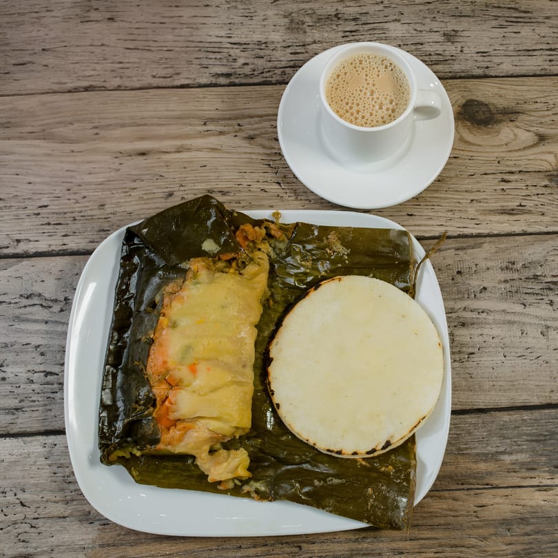 A Colombian tamal unwrapped on a white plate with an arepa and a cup of coffee.